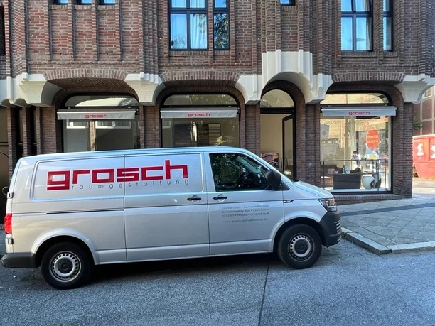 Grosch7.jpg Firmenfahrzeug der "Grosch Raumgestaltung", das vor einem Ladenlokal mit großen Bogenfenstern geparkt ist.