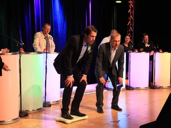 WeQuiz. Das Quizevent. Skispringen Ein Quiz-Wettbewerb mit zwei Männern im Anzug konzentriert auf Buzzer auf der Bühne.A quiz competition with two men in suits concentrating on buzzers on stage.