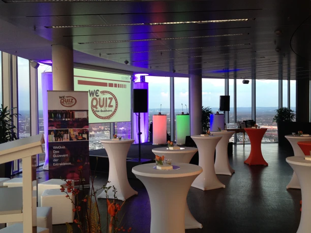 WeQuiz. Das Quizevent. Mit Ausblick Stylischer Veranstaltungsraum mit hohen Fenstern, Stehtischen und bunter Beleuchtung.</p>