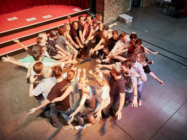 Westar. Zeigen was in uns steckt. Menschen bilden im Kreis stehend eine dynamische Pose mit ausgestreckten Armen auf einer Bühne.