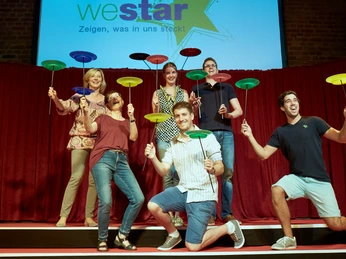 Westar. Zeigen was in uns steckt. Jonglage Junge Erwachsene balancieren bunte Teller auf Stöcken, vor einem roten Vorhang und blauem Logo.Young adults balance colorful plates on sticks in front of a red curtain and blue logo.