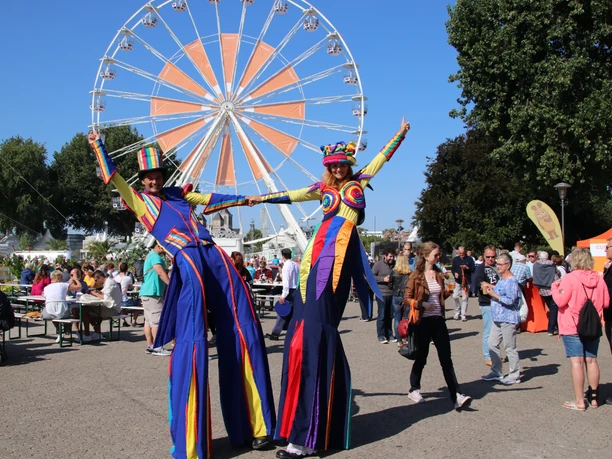 Currenta Mitarbeiterevent Riesiges Riesenrad, bunte Stelzenläufer und fröhliche Besucher bei sonnigem Fest in lebendiger Atmosphäre.