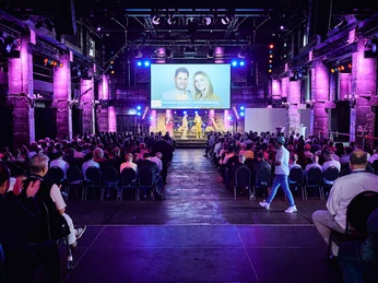 expleo Tagung Besucher in einem beleuchteten Konferenzsaal, Bühne mit Podiumsdiskussion und großem Bildschirm.</p>Visitors in an illuminated conference hall, stage with panel discussion and large screen.</p> <p