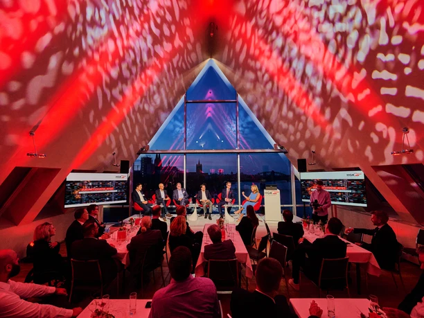 HGK Tagung Im Bild ist eine Podiumsdiskussion in einem modern beleuchteten Raum mit dreieckigen Glasfenstern.