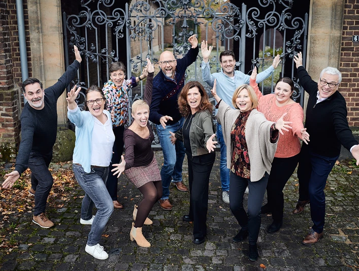 welcome Team Gruppe fröhlicher Menschen posiert vor einem schmiedeeisernen Tor mit offenherzigen Gesten.</p>Group of cheerful people pose in front of a wrought iron gate with open-hearted gestures.</p> <p