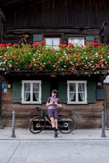 Verschnaufpause beim Gravelbiken Radfahrer lehnt an seinem Fahrrad vor einem alpenländischen Haus mit blumengeschmücktem Balkon.