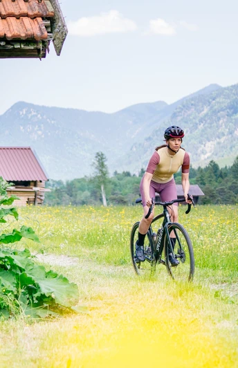 Eine Gravelbikerin fährt über einen Feldweg inmitten von Wiesen und Bergen durch die malerische Zugspitz Region.