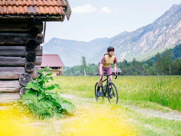 Eine Gravelbikerin fährt über einen Feldweg inmitten von Wiesen und Bergen durch die malerische Zugspitz Region.