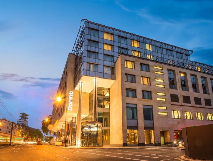 Dorint Hotel am Heumarkt Modernes Hotel mit beleuchteter Fassade bei Dämmerung, Straßenbahn fährt vorbei.</p>Modern hotel with illuminated façade at dusk, streetcar passes by</p>.