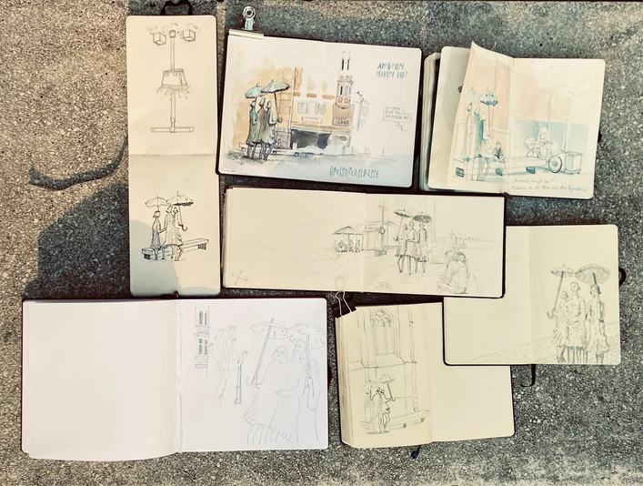 Urban Sketchers Mehrere geöffnete Skizzenbücher auf dem Boden zeigen unterschiedlich detaillierte urbane Zeichnungen von Straßenszenen mit Menschen und Architektur.