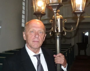 LAK Lili Marleen: Hero Boomgaarden Ein Mann in Anzug hält eine historische Straßenlaterne in einem Kirchenraum mit warmer Beleuchtung.