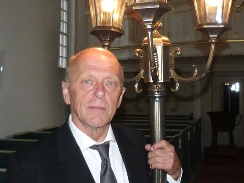 LAK Lili Marleen: Hero Boomgaarden Ein Mann in Anzug hält eine historische Straßenlaterne in einem Kirchenraum mit warmer Beleuchtung.