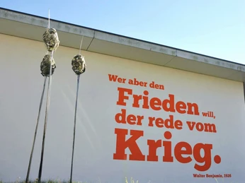 01_DPM_Benjamin-Zitat-min.jpg Weiße Gebäudefassade mit rotem Zitat: "Wer aber den Frieden will, der rede vom Krieg" (Walter Benjamin, 1926). Im Vordergrund abstrakte Skulpturen.