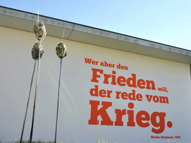 01_DPM_Benjamin-Zitat-min.jpg Weiße Gebäudefassade mit rotem Zitat: "Wer aber den Frieden will, der rede vom Krieg" (Walter Benjamin, 1926). Im Vordergrund abstrakte Skulpturen.