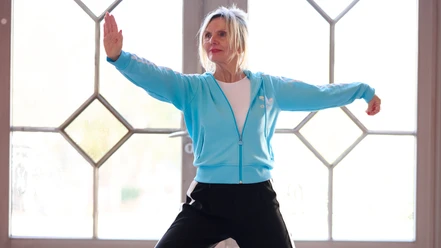 Qigong Cordula_Theine.jpg Eine Frau in blauer Jacke führt Qigong-Übungen in einem lichtdurchfluteten Raum aus.