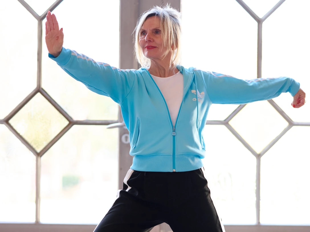 Qigong Cordula_Theine.jpg Eine Frau in blauer Jacke führt Qigong-Übungen in einem lichtdurchfluteten Raum aus.