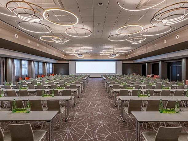 Forum 2 Parlamentarisch Ein moderner Konferenzraum mit Reihen bestuhlter Tische und einer großen Leinwand im Hintergrund.</p>