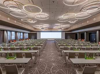 Forum 2 Schoolroom Ein moderner Konferenzraum mit Reihen bestuhlter Tische und einer großen Leinwand im Hintergrund.</p>A modern conference room with rows of tables and a large screen in the background.</p> <p