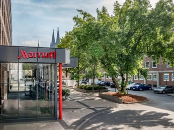 Exterior Marriott-Hoteleingang mit Glasfassade, Baum im Vordergrund und Kölner Dom im Hintergrund.Marriott hotel entrance with glass façade, tree in the foreground and Cologne Cathedral in the background.