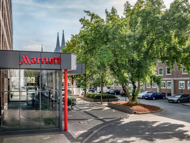 Exterior Marriott hotel entrance with glass façade, tree in the foreground and Cologne Cathedral in the background.