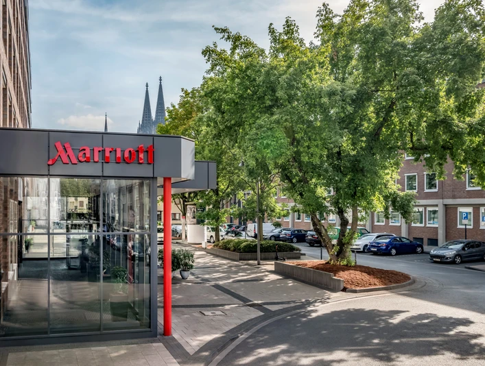 Exterior Marriott-Hoteleingang mit Glasfassade, Baum im Vordergrund und Kölner Dom im Hintergrund.Marriott hotel entrance with glass façade, tree in the foreground and Cologne Cathedral in the background.