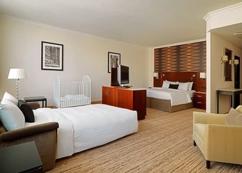 Familienzimmer 2 Helles Hotelzimmer mit zwei Betten, Babybett, Sessel, Lampe, Fernseher und moderner Wandgestaltung.</p>