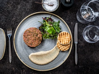 Cast Iron Grill Tartar Edles Rindertartar, Blattsalat, Creme und knuspriges Brot kunstvoll auf grauem Teller angerichtet.</p>Fine beef tartare, leaf salad, cream and crispy bread artfully arranged on a gray plate.</p> <p