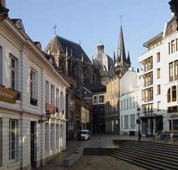 Blick auf den Aachener Dom und den Hof unter blauem Himmel durch einen Torbogen hindurch.