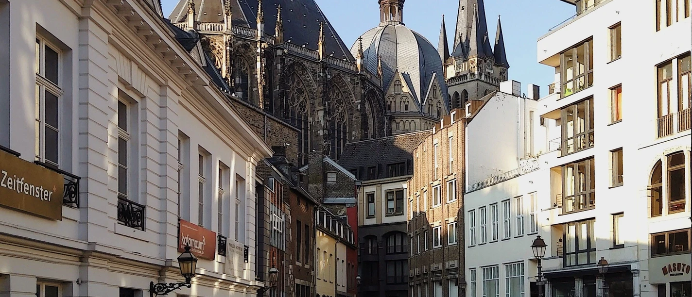 Blick auf den Aachener Dom und den Hof unter blauem Himmel durch einen Torbogen hindurch.