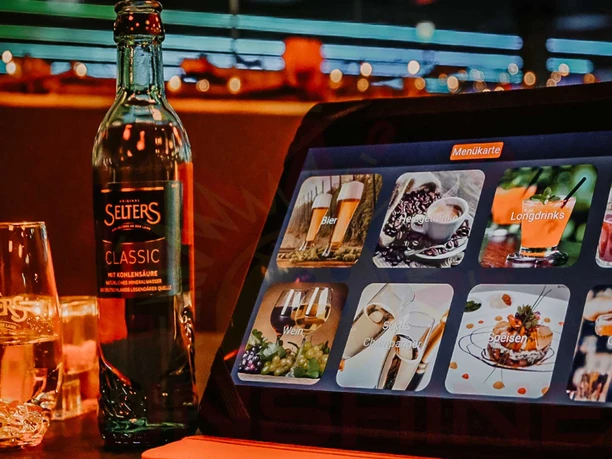 Im Restaurant des Sunshine Bowling Pirna Eine Selters Classic Flasche und ein Glas stehen neben einem Tablet, das eine digitale Getränkekarte mit verschiedenen Kategorien zeigt.