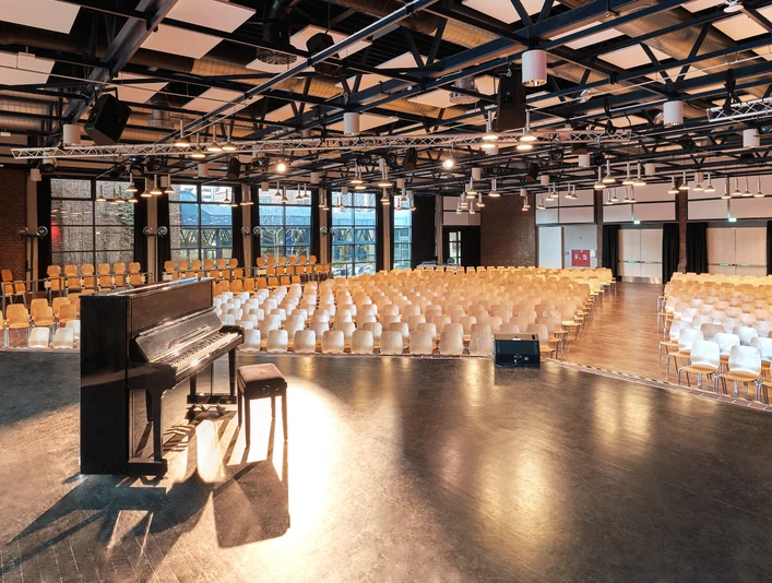 Großer Saal Großer, leerer Veranstaltungssaal mit Holzstühlen und einem schwarzen Klavier auf der Bühne.</p>Large, empty event hall with wooden chairs and a black piano on the stage.</p> <p