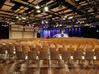 Großer Saal mit Kabarett/Musikbestuhlung Ein leerer Veranstaltungssaal mit vielen hölzernen Stühlen und bunten Lichtern auf der Bühne.An empty event hall with lots of wooden chairs and colorful lights on the stage.
