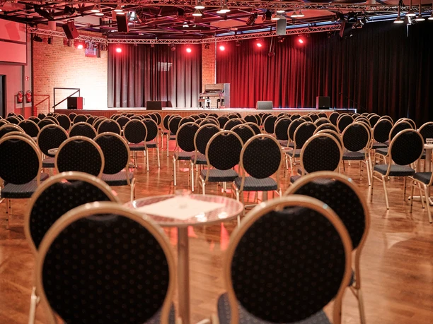 Bistrobestuhlung Großer Saal Bestuhlter Veranstaltungssaal mit Bühne, rotem Licht und Klavier, bereit für eine Aufführung.</p>
