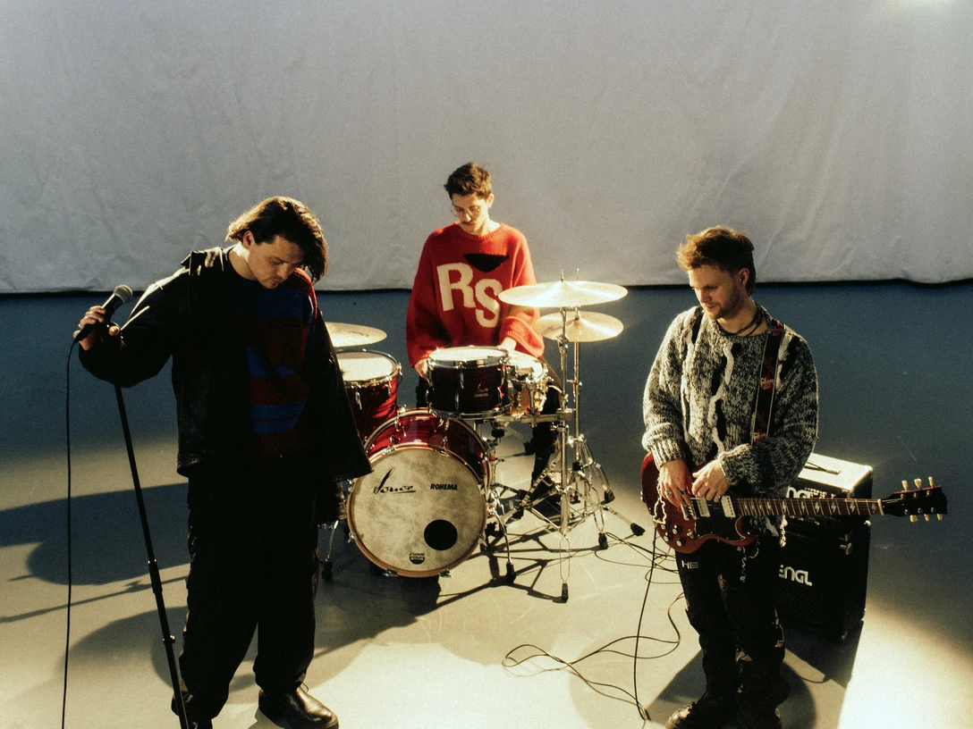 SPIRITOFMAXIM-SLINBAKER-3-EBENEZER_ABEBE.jpg Drei junge Musiker proben in einem Studio: ein Sänger am Mikrofon, ein Drummer, und ein Gitarrist.