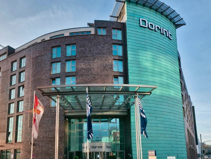 Dorint Hotel an der Messe Köln Modernes Hotelgebäude mit Glasfassade und zwei Flaggen im Vordergrund.</p>Modern hotel building with glass façade and two flags in the foreground.</p> <p