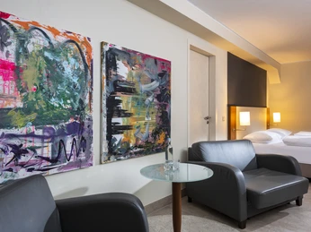 Superior Zimmer Modernes Hotelzimmer mit abstrakten Kunstwerken, zwei Sesseln und einem Glas- und Marmortisch.</p>Modern hotel room with abstract artwork, two armchairs and a glass and marble table.</p> <p