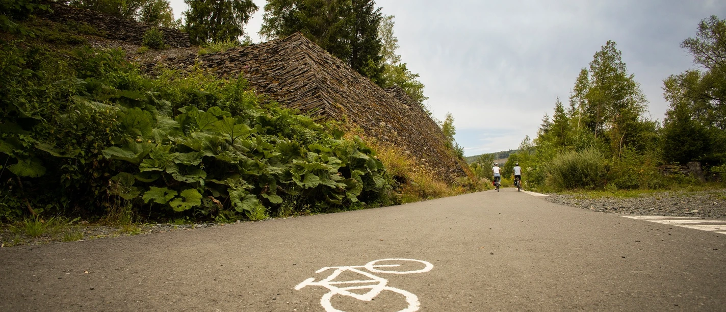 Radweg mit Fahrradsymbol, zwei Radfahrende neben Schieferhalden und Wald