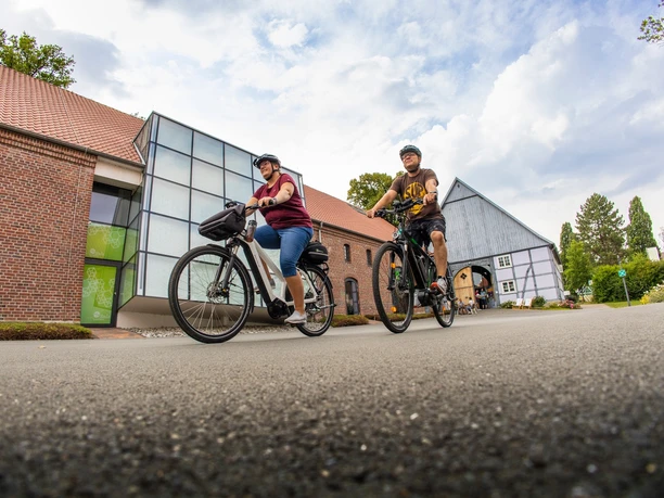 Zwei Radfahrende fahren über den Hof Haulle vor Backstein- und Fachwerkgebäuden.