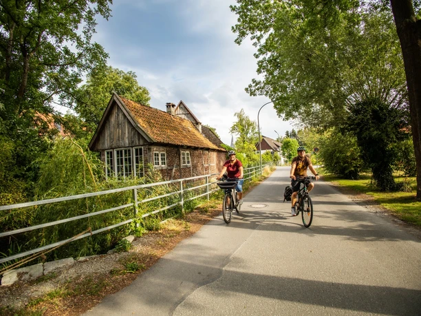 Zwei Radfahrer auf ruhiger Dorfstraße in Lohne, vorbei an altem Fachwerkhaus und Bäumen