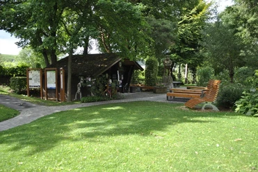 Grüne Anlage mit Holzhütte, Informationstafeln, Sitzbänken und einer Statue unter Bäumen.