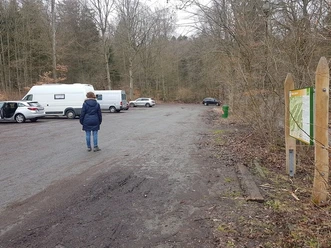 Wanderparkplatz im Wald mit mehreren Fahrzeugen, einer gehenden Person und einer Informationstafel.