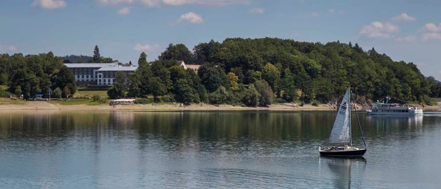 Segelboot auf dem Hennesee vor bewaldetem Ufer bei Meschede unter wolkigem Himmel