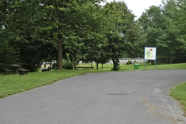 Asphaltierte Parkfläche mit Bäumen, Sitzbänken, Übersichtstafel und Abfalleimer.