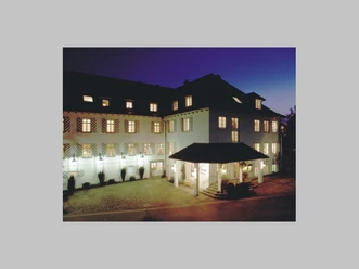 Beleuchtetes, weißes Hotelgebäude mit gestreiften Fensterläden am Abend.