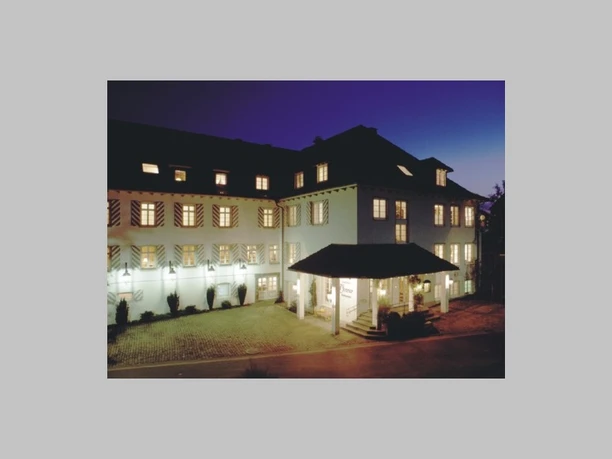 Beleuchtetes, weißes Hotelgebäude mit gestreiften Fensterläden am Abend.