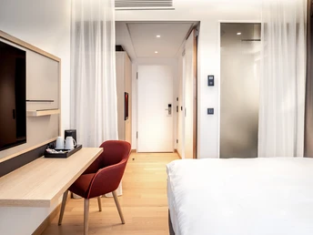 Modernes Hotelzimmer mit hellem Holzfußboden, rotem Stuhl, Schreibtisch, Flachbild-TV und Doppelbett.