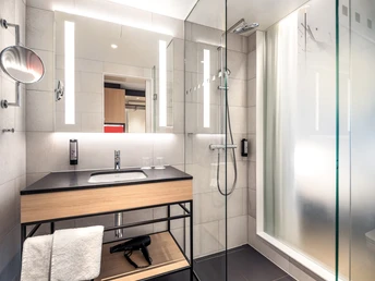 Modernes Hotelzimmerbad mit Glasdusche, großem Spiegel, hellem Waschbecken und Föhn.