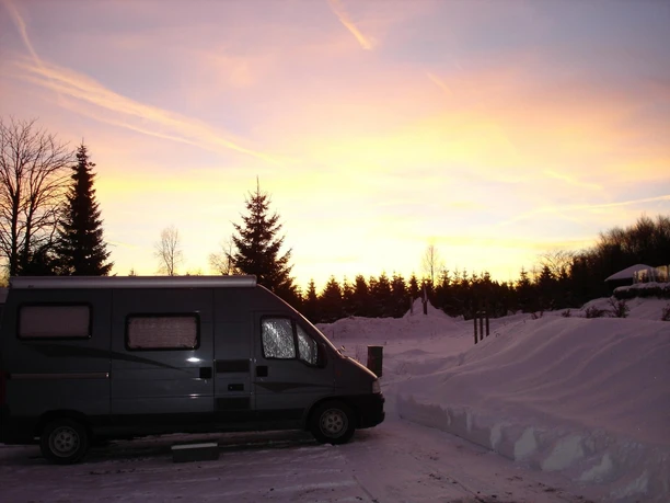 Wohnmobil auf verschneitem Stellplatz vor Wald im Abendlicht