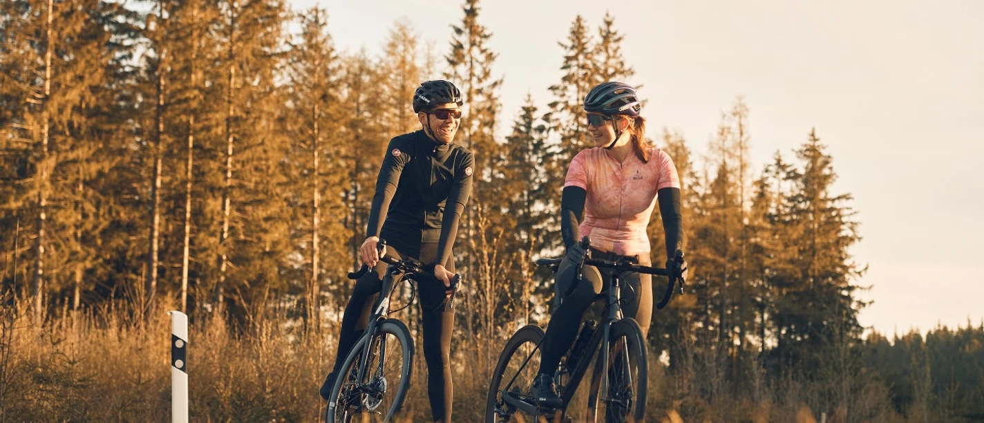 Zwei Rennradfahrer stehen im warmen Abendlicht vor Nadelwald.