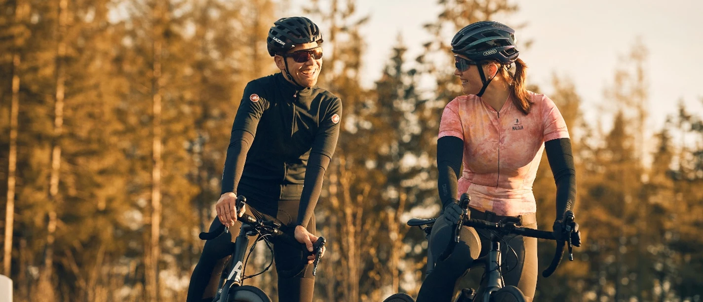 Zwei Rennradfahrer mit Helmen lachen in der Abendsonne vor herbstlichem Wald